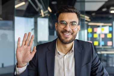Modern ofis ortamında kendine güvenen bir işadamı, doğrudan kameraya bakarak bir video görüşmesine katılıyor. El sallar, profesyonellik ve dostluk taslar, takım elbise giyer.