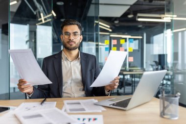 Modern bir ofiste evrakları inceleyen bir iş adamı. Masasında bir dizüstü bilgisayar ve kağıtlarla, kurumsal bir ortamda odak ve verimliliği temsil ediyor. Ayarlar profesyonel analizi teşvik ediyor..