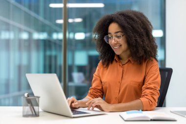 Modern bir ofiste dizüstü bilgisayarla çalışırken gülümseyen genç bir iş kadını. Odaklanması ve eğlenmesi verimli bir çalışma alanı atmosferini vurguluyor. İş yeri, teknoloji ve mutluluk kavramları için ideal.