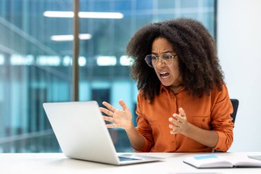 Genç iş kadını dizüstü bilgisayarına bakarken hayal kırıklığını ifade ediyor. Kızgın ve stresli görünüyor, elleriyle işaret ediyor. Modern ofis ortamı işle ilgili bir sorun öneriyor.