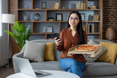 Asyalı kadın evde pizza ve telefonla dinleniyor. Oturma odasında dizüstü bilgisayarla kanepede oturmak. Modern dekorlu ve boş zaman için rahat bir hafta sonu havası..