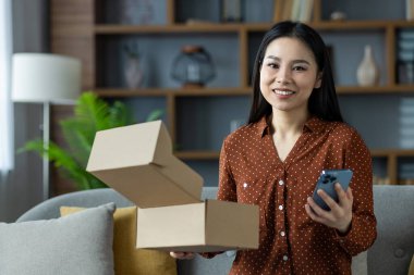 Evinde telefon ve karton kutular tutan mutlu Asyalı kadın modern teknoloji ve kolaylığı gösteriyor. E-ticaretin ve online alışveriş deneyiminin özünü yakalar.