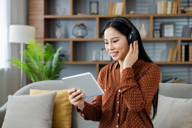 Evde kulaklıkla müzik dinleyen Asyalı bir kadın, rahat oturma odasındaki kanepede oturmuş tablet kullanıyordu. Sahne gevşemeyi, teknolojiyi ve günlük eğlenceyi betimler..