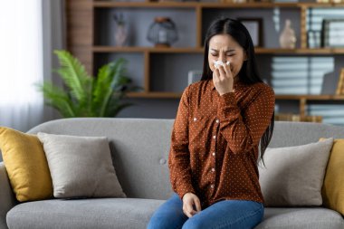 Asyalı kadın oturma odasında kanepede oturuyor, burnunu mendille siliyor. Soğuk algınlığı ya da alerjisi var gibi görünüyor. Sağlık ve sağlık sorunlarını yansıtıyor..