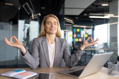 Olgun bir iş kadını modern ofis ortamında masasında meditasyon yapıyor. Sakin ve odaklanmış görünüyor. İş yerindeki farkındalığın ve stres yönetiminin önemini vurguluyor..