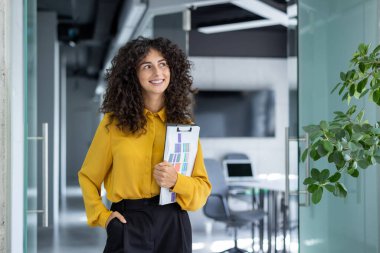 Modern ofis ortamında belgeleri elinde tutan kendine güvenen bir iş kadını. İfadesi, kurumsal ve profesyonel temalar için ideal olumluluk ve hazırlık içeriyor..