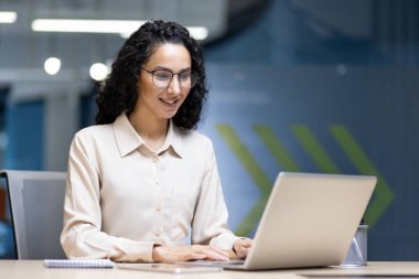 Kendine güvenen bir iş kadını modern bir ofiste dizüstü bilgisayarında yazı yazıyor. Gülümsemesi ve odaklanmış ifadesi verimliliği ve bağlılığı gösteriyor. Bu iş ortamı profesyonelliği yansıtıyor