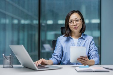 Asyalı bir iş kadını, modern bir ofisteki masasında tablet ve dizüstü bilgisayarla etkili bir şekilde çalışıyor. Odaklanmış ifadesi verimliliği ve profesyonelliği gösteriyor..