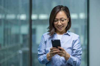 Modern ofis ortamında Asyalı bir kadın akıllı telefonuna bakarken gülümsüyor. İş kıyafetleri ve olumlu tavırları profesyonel bir atmosfer olduğunu gösteriyor..