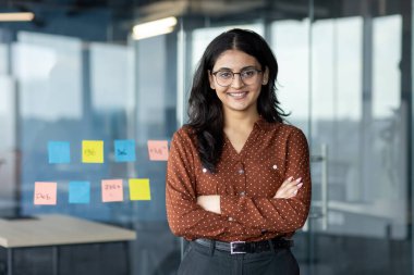 Kendine güvenen genç bir iş kadını modern ofis ortamında kollarını kavuşturmuş duruyor. Gözlük takıyor ve gülümsüyor. Profesyonelliği ve liderliği simgeliyor..