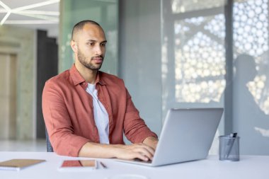 Kendine güvenen işadamı, parlak ve modern bir ofiste özenle dizüstü bilgisayar üzerinde çalışıyor. Profesyonel ayarlar, bir defter, kalem ve tablet gibi temel gereksinimleri içerir, odaklanmayı ve üretkenliği vurgular.