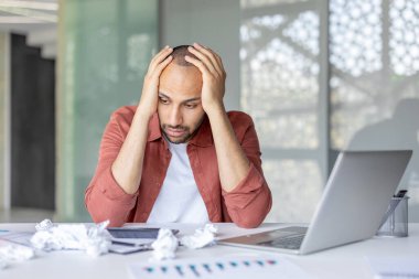 Bir ofis çalışanı masasında otururken hayal kırıklığına uğramış görünür, buruşmuş kağıtlar ve bir dizüstü bilgisayarla çevrili. Stres ve endişenin ifadesi çok açık..