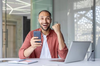 Neşeli bir adam modern bir ofiste akıllı telefon tutarak zaferi kutlar. Masada başarıyı, heyecanı ve dijital başarıyı simgeleyen bir dizüstü bilgisayarı var..