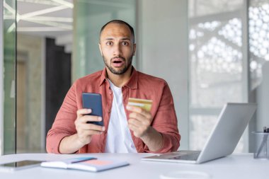 Şaşırmış bir adam masada otururken elinde akıllı telefon ve kredi kartı tutuyor. Şok olmuş görünüyor, muhtemelen beklenmedik haberler ya da online alışveriş sorunları yüzünden..