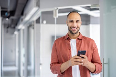 Genç bir adam modern bir ofiste kendine güvenerek duruyor, akıllı telefonunu kullanarak gülümsüyor. Günlük kıyafetleri profesyonel arka planla çelişiyor. Rahatlamış ama üretken bir atmosfer yaratıyor..