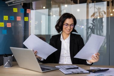 Ofis masasında dizüstü bilgisayarla belgeleri analiz eden bir iş kadını. Modern çalışma alanındaki profesyonel kadın kağıtları inceliyor ve gülümsüyor, odaklanıyor ve kendine güveniyor..