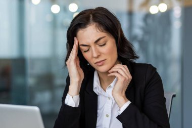Ofisteki iş kadını stresli hissediyor, el ele tutuşuyor, iş yerinde baskı ve yorgunluk belirtileri gösteriyor. Görüntü, modern bir iş yerinde hayal kırıklığı ve endişe duygularını yansıtır..