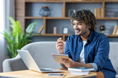 Genç bir adam internette öğrenmekle meşgul, rahat bir koltukta oturuyor elinde dizüstü bilgisayarı ve elinde bir defterle. Çalışmaları için dijital aletler kullanarak odaklanmış ve hevesli görünüyor..