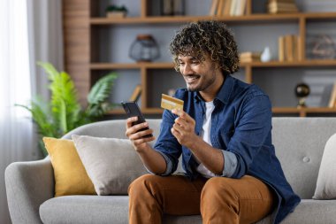Kanepede oturan mutlu bir adam telefonunu ve kredi kartını online alışveriş için kullanıyor. Bu görüntü, rahat bir ev ortamında kolaylık, teknoloji ve modern tüketiciliği yakalar..
