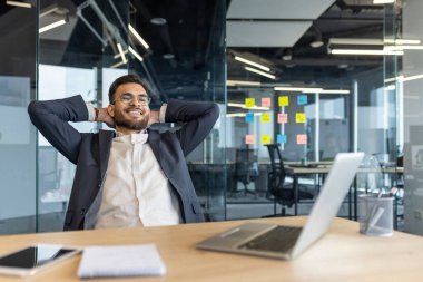 Gülümseyen bir işadamı arkasına yaslanır, modern bir ofiste masasında rahat hisseder. Etrafı cam duvarlarla ve teknolojiyle çevrili, profesyonel ortamında güven ve memnuniyet yayıyor..