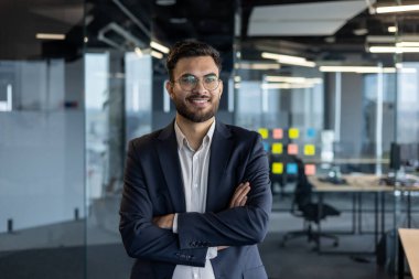 Profesyonel bir işadamı, cam duvarları ve parlak ışıklandırması olan, pozitif enerji ve başarı saçan modern bir ofis ortamında kendine güvenerek duruyor..