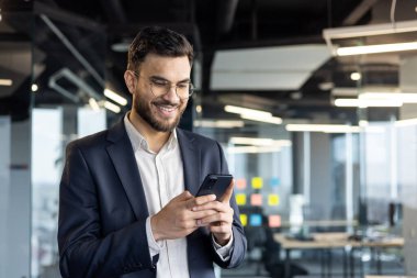 Takım elbiseli genç bir işadamı modern ofis ortamında akıllı telefon kullanarak gülümsüyor. Görüntü profesyonelliği, teknoloji kullanımını ve olumlu bir iş ortamını aktarıyor.