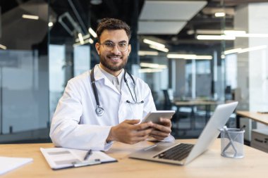 Bir doktor modern ofis ortamında tablet ve laptop kullanırken gülümser. Profesyonel atmosfer ve teknolojinin kullanımı sağlık ortamlarında verimliliği ve bağlantıyı vurguluyor.