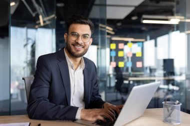 Takım elbiseli kendine güvenen bir işadamı, parlak ve modern bir ofiste dizüstü bilgisayar üzerinde çalışıyor. Ofiste cam duvarlar ve renkli yapışkan notlar yer alıyor, açık ve işbirlikçi bir çalışma ortamı yaratıyor.