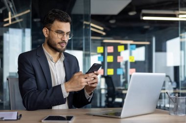 Modern ofis ortamında dizüstü bilgisayarlı bir masada otururken akıllı telefon kullanan odaklanmış bir iş adamı. Görüntü, teknoloji ve iletişim araçlarıyla profesyonel bir ayar yakalıyor.