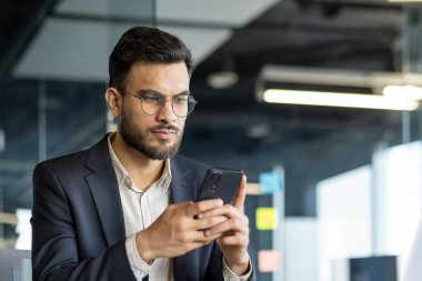 Resmi takım elbiseli bir işadamı akıllı telefonuna dikkatle bakar ve bir işe odaklanır. Modern ofis ortamı, üretkenlik ve konsantrasyonun resmedildiği sahneye profesyonel bir ortam ekler..