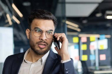 Takım elbiseli profesyonel bir adam akıllı telefondan iletişim kurar, modern ofis ortamında odak ve endişeyi ifade eder. Yoğun ifadesi önemli bir sohbete katıldığını gösteriyor..