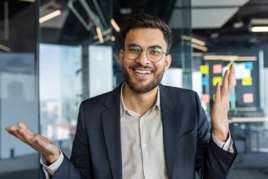 Takım elbiseli, gülümseyen ve el kol hareketi yapan kendine güvenen bir adam modern ofis ortamında bir video görüşmesinde profesyonelliği ve nişanı iletiyor..