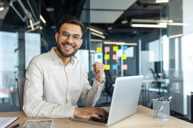 Neşeli bir erkek ofis çalışanı çalışma başarısını masasında dizüstü bilgisayarla kutluyor. Gülümsüyor ve heyecanlı görünüyor. Arka planda renkli yapışkan notlar var..
