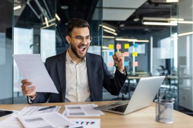 Takım elbiseli neşeli bir işadamı ofis masasında başarılı bir başarıyı kutluyor. Resim, bir dizüstü bilgisayardaki belgeleri incelerken bir neşe ve motivasyon anı yakalıyor.
