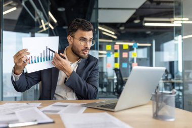 Resmi takım elbiseli bir işadamı, modern ofisinden online bir toplantıda harita verilerini sunuyor. Bir şirket ortamında güven ve profesyonellik sergilemek için bir kalem kullanıyor..