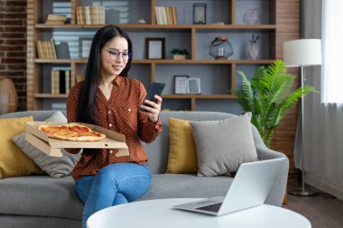 Pizzayı seven Asyalı bir kadın, evdeki koltukta otururken akıllı telefon kullanıyor. Modern yaşam tarzı teknolojiyi kucaklıyor. Laptop, telefon ve günlük kıyafetlerle rahat, rahat bir an..