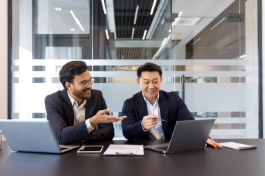 Çağdaş bir ofis ortamında iki işadamı ortak bir toplantıda. Dizüstü bilgisayarlar ve belgeler kullanarak iş planlaması için dinamik ve profesyonel bir atmosfer yaratıyorlar.