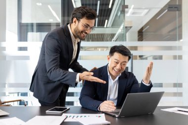 İki işadamı modern ofis ortamında bir başarıyı sevinçle kutluyorlar. Biri dizüstü bilgisayar kullanırken, diğeri heyecanla işaret ediyor. İfadeleri mutluluk ve takım çalışmasını gösteriyor..