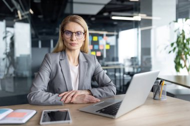 Profesyonel bir iş kadını, teknoloji ve profesyonelliğin bir karışımını yansıtan bir dizüstü bilgisayar ve akıllı telefonla donatılmış modern bir ofis ortamında güvenle masasına oturur..