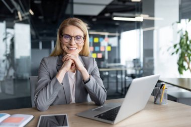 Profesyonel ortamdaki kendine güvenen bir iş kadını, elinde dizüstü bilgisayar ve akıllı telefonla masasında oturuyor, kameraya gülümsüyor, işyerindeki başarıyı ve profesyonelliği simgeliyor..