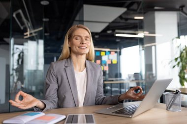 Gri takım elbiseli bir iş kadını ofis masasında farkındalık meditasyonu yapıyor yoğun bir iş gününde rahatlama ve stresi arttırıyor. O, işi ve refahı etkin bir şekilde dengeliyor..