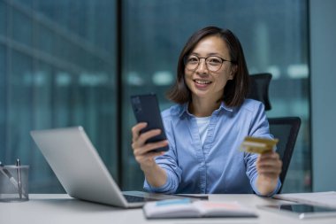 Çevrimiçi bankacılık için akıllı telefon ve kredi kartı kullanırken gülümseyen Asyalı iş kadını. Ofis masasında dizüstü bilgisayarla oturuyor, profesyonel ve kendinden emin bir tavır sergiliyor..