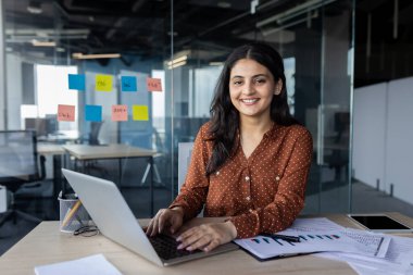 Modern ofis ortamında dizüstü bilgisayarda çalışan profesyonel bir iş kadını. Noktalı bir gömlek giyiyordu, masasında oturuyordu. Etrafı belgelerle çevriliydi, kendine güveni ve profesyonelliği vardı..