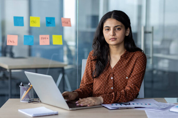 A confident businesswoman sitting at her desk using a laptop in a professional environment, focusing on her tasks. The office setting suggests organization and productivity as she proceeds with work.