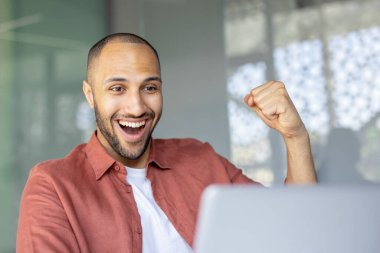 Neşeli bir ifadeyle gülümseyen genç adam ofis ortamında dizüstü bilgisayarı üzerinde çalışırken başarıyı kutluyor. Modern bir çalışma alanında başarı, mutluluk ve profesyonel başarı kavramı.