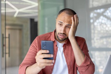 Ofisteki bir adam endişeli bir ifadeyle telefonuna bakar. Endişe, hayal kırıklığı ya da eleştirel bir düşünceyle. Resim, işle ya da kişisel iletişimle bağlantılı duygusal stresi betimliyor.