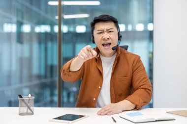 Olgun Asyalı işadamı, kulaklık kullanarak konuşurken hayal kırıklığını ifade ediyor. Dinamik, haplar ve güven gibi teknolojinin yer aldığı bir ofis ortamında yoğun bir iletişimi iletir.