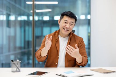 Bir ofis ortamında masa başında otururken fikirleri ifade eden Asyalı bir adam, ticari bir tartışma veya toplantı sırasında güven, olumluluk ve iletişimi iletiyor.