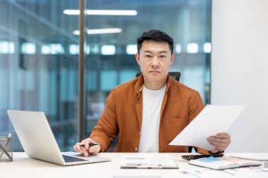 Olgun bir Asyalı, belgeleri inceliyor ve modern bir ofisteki bir masada dizüstü bilgisayarla çalışıyor. Bir iş ortamında üretkenlik, profesyonellik ve teknolojiyi temsil eder.