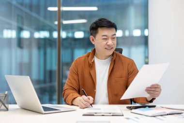 Asyalı bir adam masasında dizüstü bilgisayarla evrakları inceliyor, profesyonel bir hava yaratıyor.